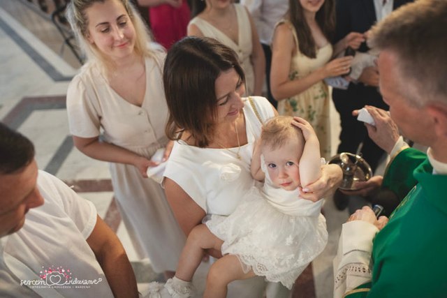 Mama trzyma na rękach małą dziewczynkę podczas ceremonii Chrztu Świętego w parafii św. Patryka na Gocławiu w Warszawie – reportaż z chrztu Sercem Widziane.